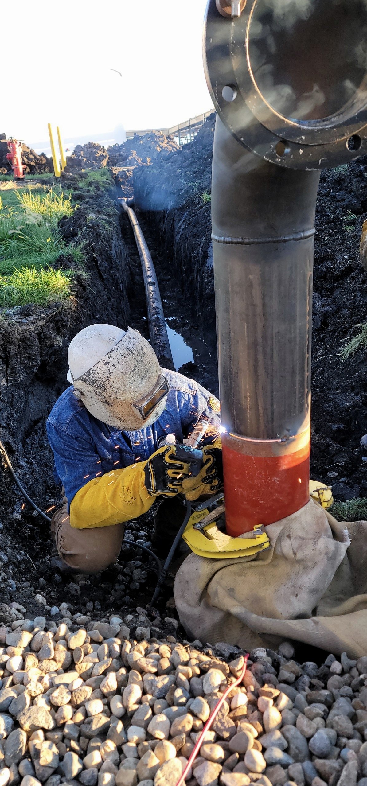 Welder in a pancake hood working a pipeline riser tie-in weld in an open trench, sparks flying